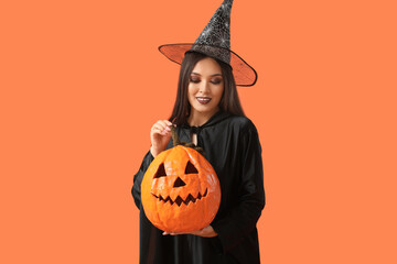 Beautiful young happy Asian woman dressed as witch for Halloween with carved pumpkin on orange...