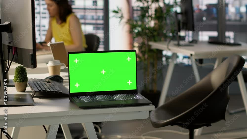 Poster green screen laptop on desk in workspace with admins in background doing software quality assurance.