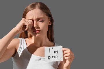 Crying young woman holding paper with text I'M FINE on dark background, closeup