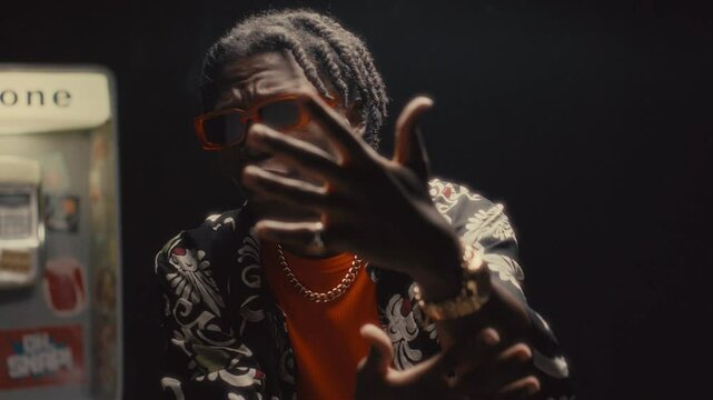 Black man wearing orange-tinted sunglasses gesturing expressively and rapping to camera in front of old-fashioned public payphone in city street at night