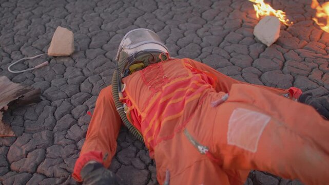 Astronaut in orange overalls resting on cracked ground with clods of soil
