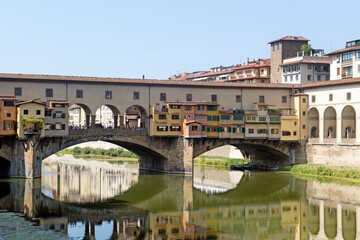 Obraz premium The historic Ponte Vecchio, the oldest bridge over the Arno River in Florence, Italy, featuring its famous shops built along the bridge and reflecting beautifully on the calm water below. 