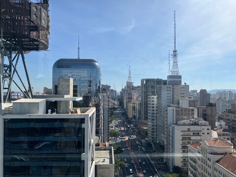 Foto da vista panor&acirc;mica da Avenida Paulista em S&atilde;o Paulo, Brasil, com arranha-c&eacute;us modernos, ruas movimentadas e a vida urbana sob um c&eacute;u azul claro. Um vibrante s&iacute;mbolo da energia da cidade.
