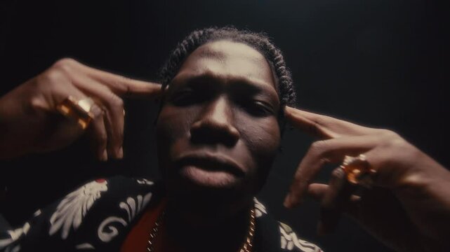 Cool-looking Black man with dreadlocks wearing stylish shirt, chain necklace and rings bobbing his head to hip-hop music with fingers pointing to his temples in studio with dark background
