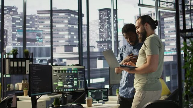 IT employees in office working together using laptop, testing and deploying programs and systems. Developer and colleague checking code on notebook, doing brainstorming in workspace, camera B