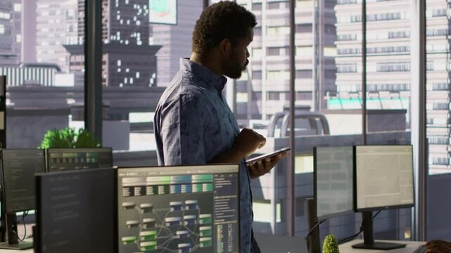Computer scientist resuming work after strolling in workspace, doing software debugging using PC. Man walking with tablet in office, fixing coding issues using computer, camera A