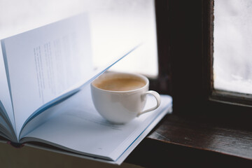 A warm cup of coffee rests beside an open book on a windowsill, while a lit candle flickers softly, creating a serene atmosphere against the snowy backdrop.