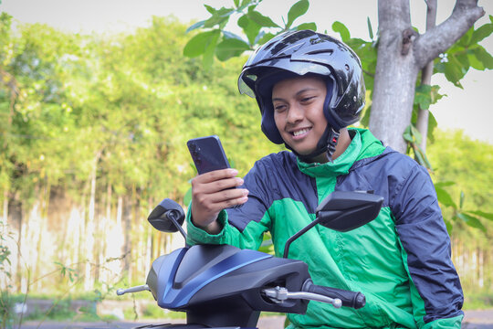 Online taxi rider wearing green jacket and helmet, riding motorcycle, using a mobile phone while on the way to pick up customer. Transportation application concept.