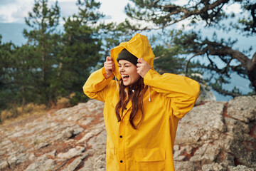 A woman in a yellow raincoat standing atop a majestic mountain with hands on head Traveler's Moment...
