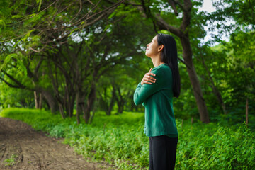 An Asian woman is standing happily in a green forest park, enjoying the fresh air and nature. She takes a deep breath, looks up at the morning sunlight, and feels relaxed and peaceful.