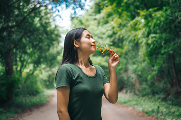 Fototapeta premium An Asian woman is happily relaxing in a green forest garden, smelling flowers and enjoying the fresh air. She takes a deep breath, looks up at the morning sunlight, and feels at peace with nature