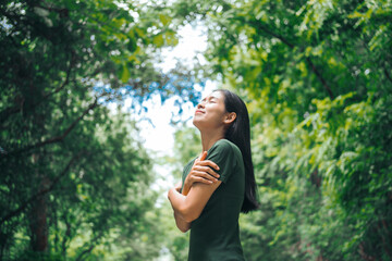 An Asian woman is standing happily in a green forest park, enjoying the fresh air and nature. She takes a deep breath, looks up at the morning sunlight, and feels relaxed and peaceful.