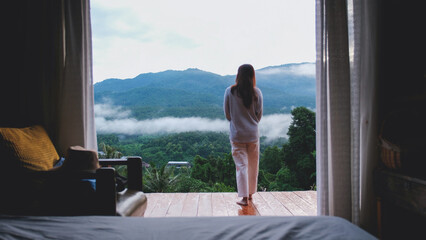 Rear view of a woman standing on wooden balcony and looking at a beautiful mountains view on foggy day