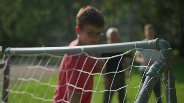 A ball is shot into a goal post as the goalkeeper in red misses the save and adjusts his glasses, with blur view of two person standing in the background