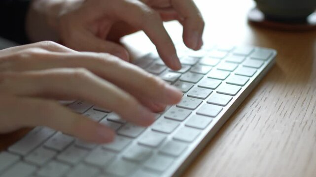 白いキーボードでタイピングしている手。木製のデスクで文章を入力したりプログラミングしたりして仕事をしているイメージ
