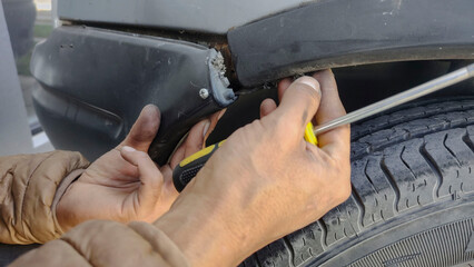 Close up of the hands of a man in a brown jacket, fixing the plastic bumper of a crashed car, with...
