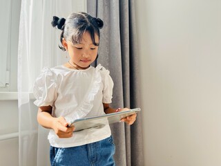Asian little girl standing near the window holding mobile tablet. Screen time, phone addictive concept