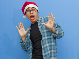 Naklejka premium Portrait of a young Asian man wearing a Santa hat, showing a funny shocked expression while looking forward, isolated against a blue background. Ideal for holiday and seasonal content.