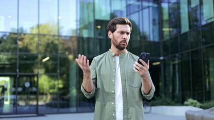 A young businessman experiences frustration as he struggles with a poor internet connection on his smartphone. Standing outside a modern office building, he gestures in exasperation,