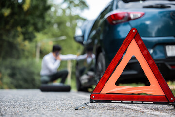 Red triangle sign placed behind a broken car on the side of the road. Businessman changing the car wheel while sitting on the roadside sign. Concept of road safety and emergency assistance.