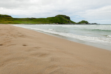 Obraz premium Gentle waves roll onto the sandy shore, set against a backdrop of rolling green hills and a dramatic sky, creating a serene coastal atmosphere.