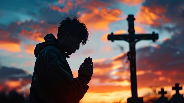 A young person praying in silhouette against a dramatic sunset in a cemetery