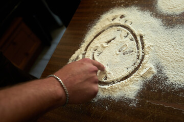 A group of people stand around a wooden table and draw pictures from flour.