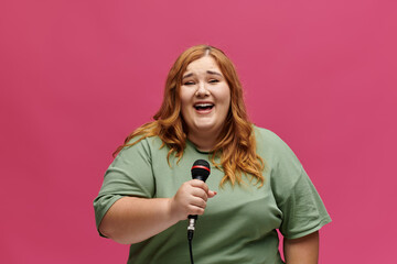 A plus size woman with red hair sings into a microphone, smiling brightly.