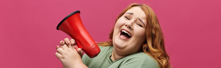 A woman with long red hair is shouting into a red megaphone with a smile on her face.