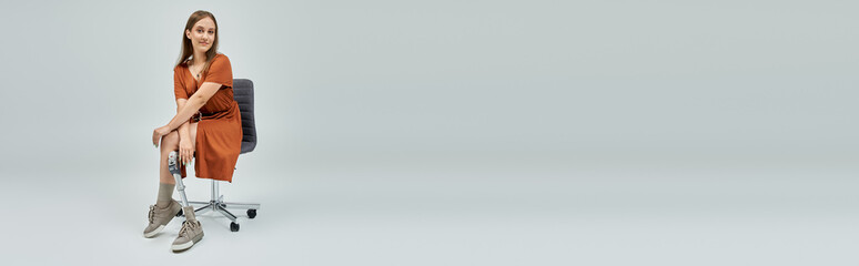 A woman with a prosthetic leg sits in a chair, wearing a rust-colored dress.