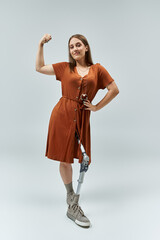 A woman in a brown dress stands with her arm raised, confidently displaying her prosthetic leg.