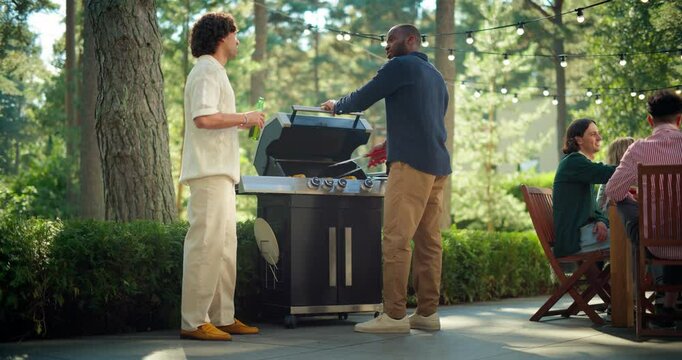 African American and Caucasian Men Grill Food Outdoors and Enjoy Drinks at a Barbecue Party. Diverse Friends Sitting Behind a Table Under String Lights in a Beautiful Green Garden with Trees