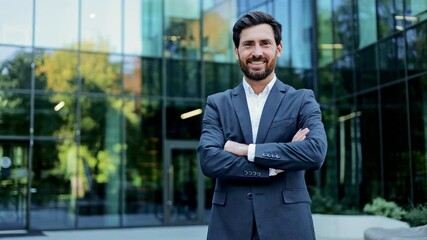 A confident and successful businessman in a suit smiles at the camera with his arms crossed in front of a modern glass office building. - Powered by Adobe
