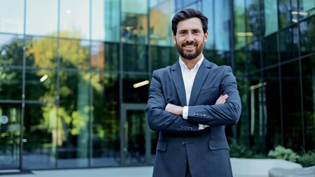 A confident and successful businessman in a suit smiles at the camera with his arms crossed in front of a modern glass office building.