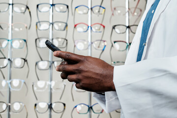 Fototapeta premium Optometrist in White Coat Using Smartphone in Eyeglass Store with Display of Stylish Spectacles