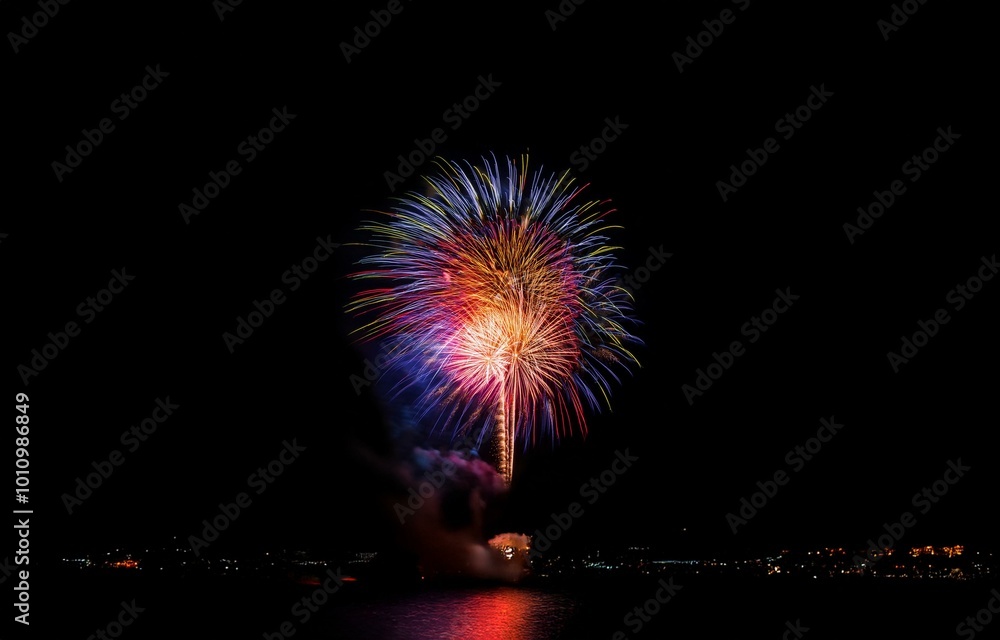 Poster a large firework display explodes over a city skyline reflecting in water.