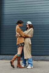 Two young women dressed in trendy outfits stand close, embracing each other in an urban setting.