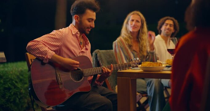 Portrait of Friends Sit Around a Table, Sharing Food and Drinks, Laughing while One of the Group Plays the Guitar. Impromptu Music Session Adds a Special Touch to Their Fun Evening Together