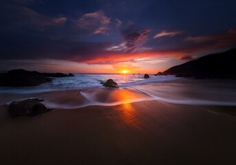 Sunset over the ocean, the sand reflecting orange hues, with dark rocks and soft waves.