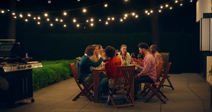 Lively Garden Party with Friends and Family Enjoying Food, Drinks, and Laughter Under String Lights Outdoors. People Raising Glasses for a Toast and Enjoy Joyful Night Atmosphere
