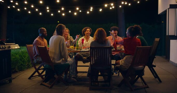 Lively Garden Party with Friends and Family Enjoying Food, Drinks, and Laughter Under String Lights Outdoors. People Raising Glasses for a Toast and Enjoy Joyful Night Atmosphere