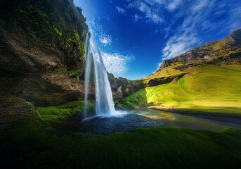 A waterfall cascades down a moss-covered cliff face into a tranquil pool, framed by a lush green valley and a clear blue sky.