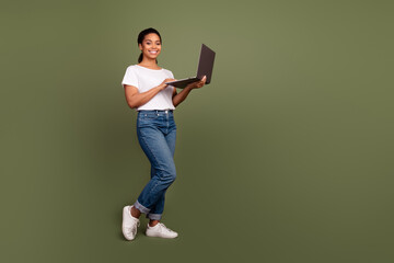 Photo portrait of cheerful young lady in jeans and t shirt working remote anywhere with netbook isolated on khaki color background