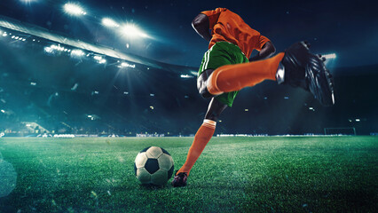 Low angle view on focused man, soccer player in orange uniform, kicking ball with precision against evening sky and fans tribune on background. Concept of sport, tournament, competition, game event