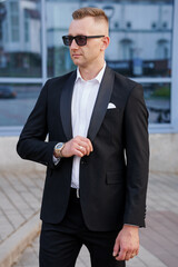 Elegant Man in Tailored Black Suit with Sunglasses and Stylish Watches Posing Outdoors