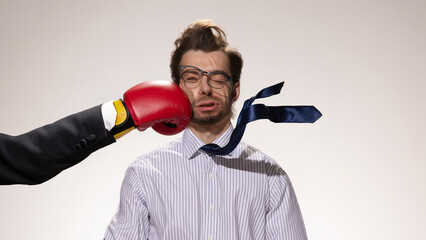 Naklejka premium Red boxing glove slams into man's face, distorting his expression as his tie and hair fly against white studio background. Concept of social media, business, negative emotions.