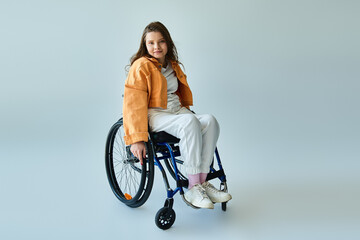 A young woman with long, brunette hair sits in a wheelchair in a studio setting with a grey...