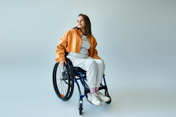 A young woman with long brown hair sits in a wheelchair in a studio setting, looking away from the...
