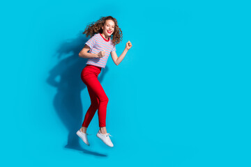 Cheerful young woman jumping joyfully in vibrant red pants and striped shirt against blue background, exuding high energy positive emotion in studio setting