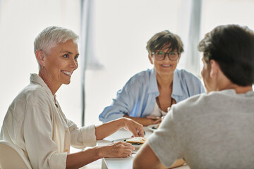 Lesbian parents share a joyful breakfast moment with their adult son at a sunlit kitchen table.